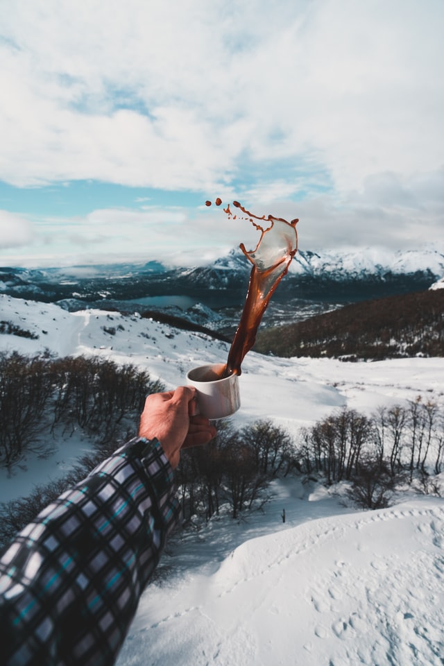 A hand holding a mug of hot chocolate that's splashing, against snowy mountains